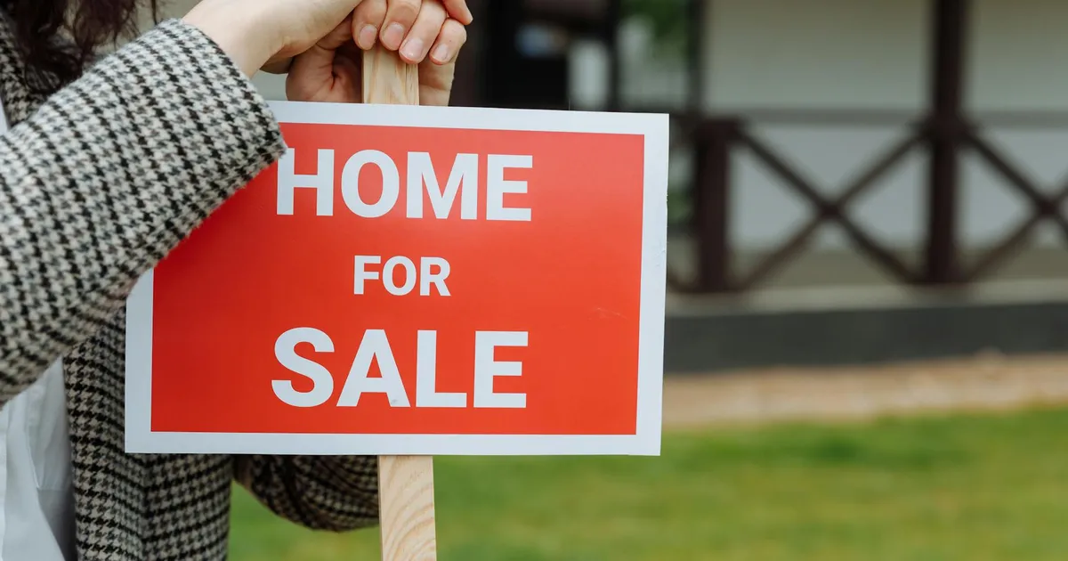 Close-up of a home for sale sign held by a person outdoors in a yard.