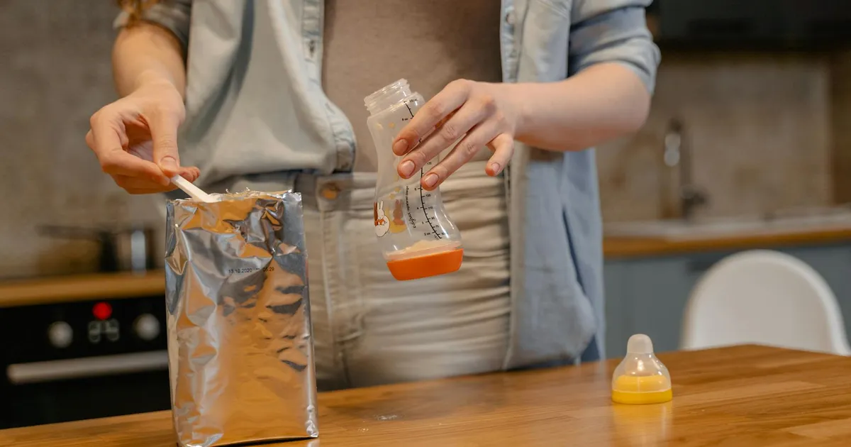 A parent prepares a baby bottle with formula on a wooden table in a cozy kitchen setting.