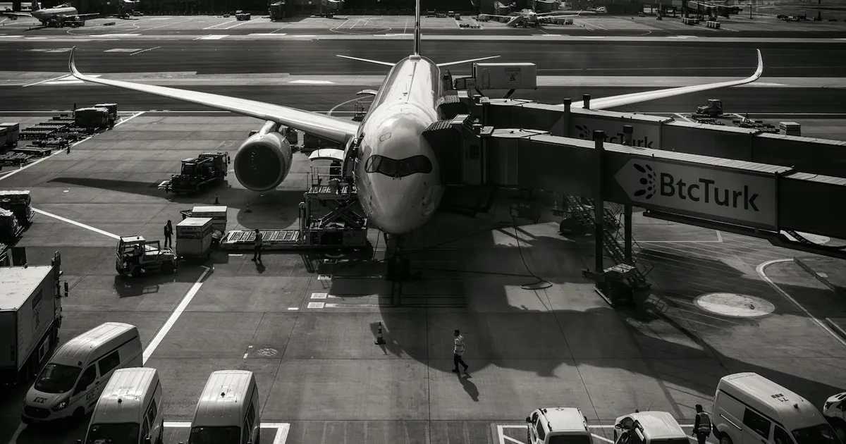 Monochrome image of airplane at airport gate with vehicles and jetway during the day.