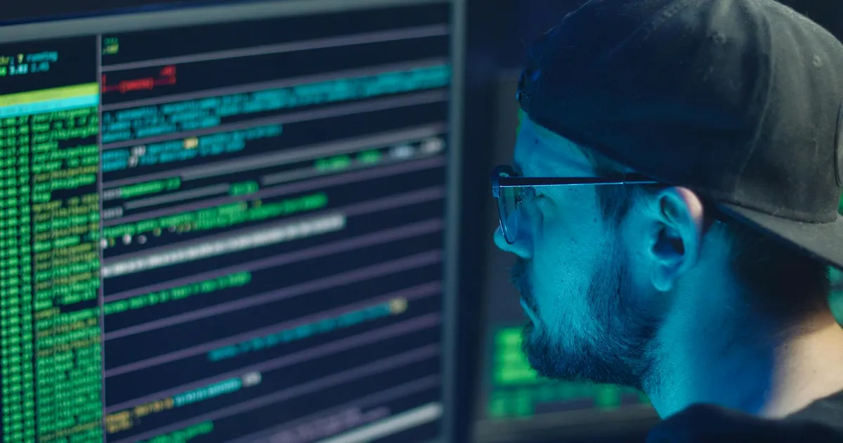 A cybersecurity expert inspecting lines of code on multiple monitors in a dimly lit office.