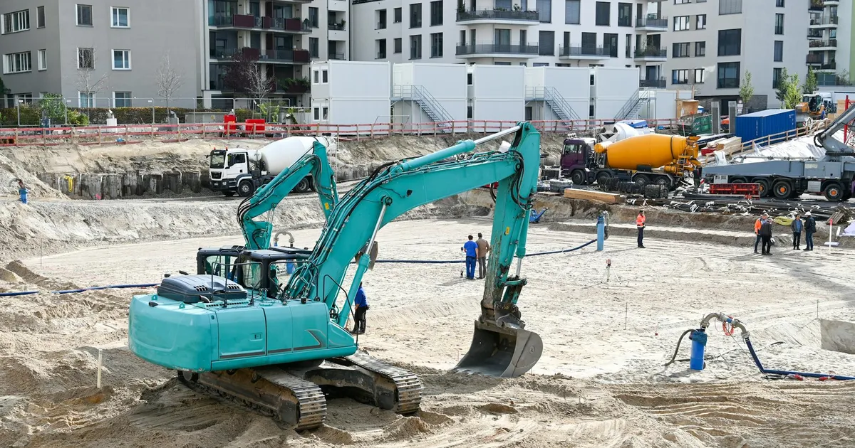 A busy construction site with machinery, workers, and residential buildings in the background.