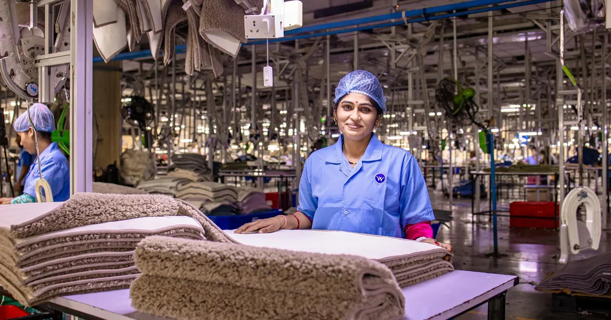 A female worker in a textile factory smiling, showcasing a stack of carpets.
