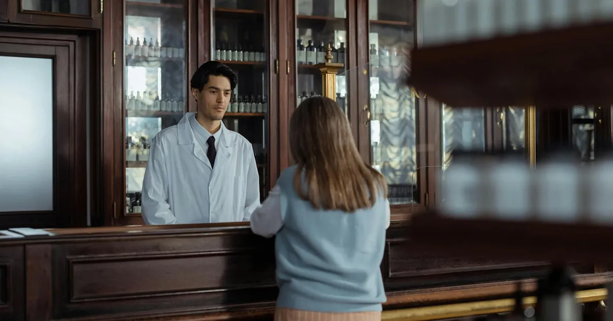 A pharmacist assists a customer at a vintage wooden pharmacy counter indoors.