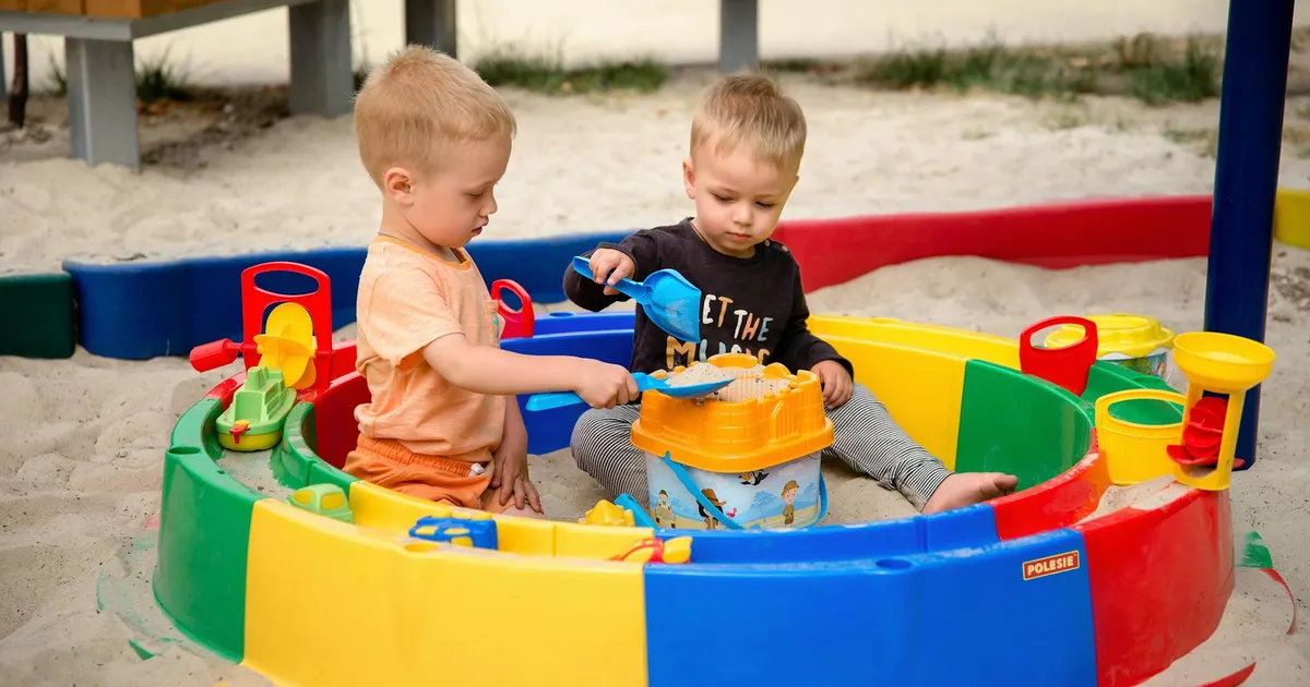 Two young children playing with toys in a colorful outdoor sandpit on a sunny day.