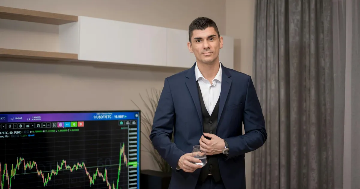 A businessman in a suit stands indoors with a stock market graph on a screen.