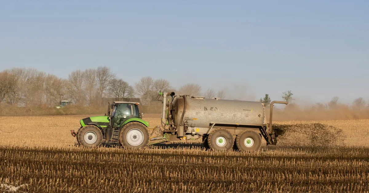 A tractor with a tanker spreading fertilizer over a barren farmland under clear skies.