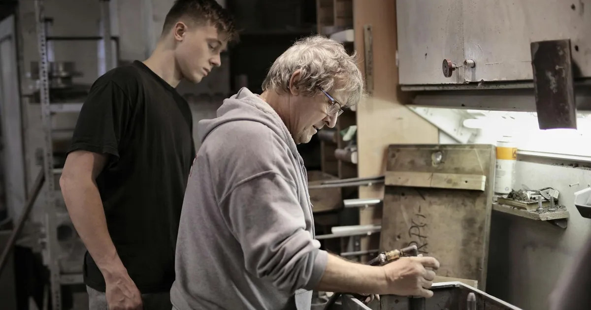 Side view of senior worker in eyeglasses and trainee handling detail on workbench in workshop