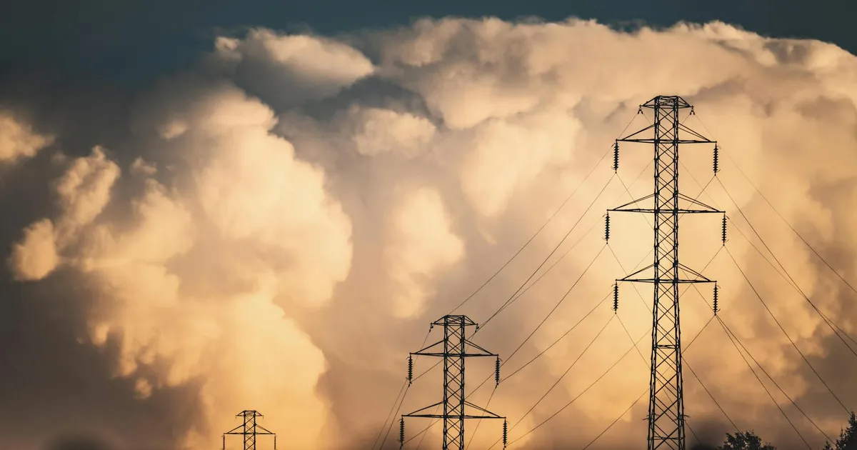 High voltage power lines and towers silhouetted against large dramatic clouds in a rural field setting.