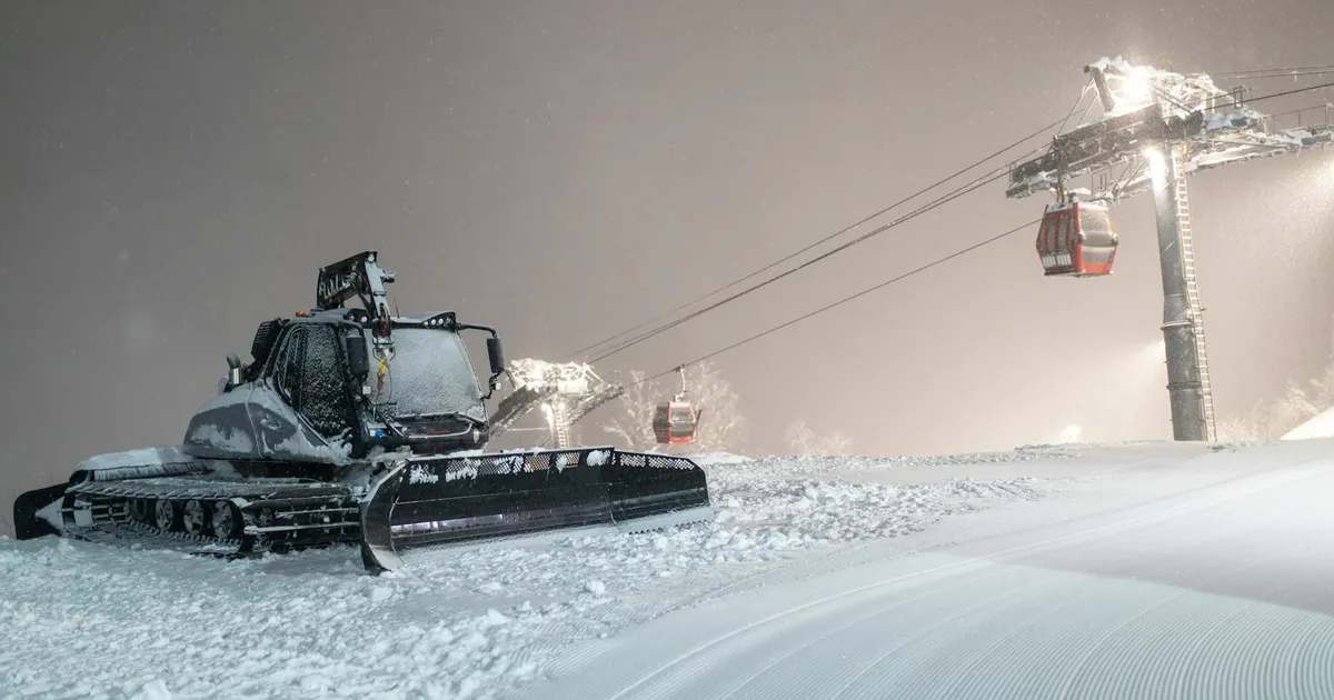 A snowcat working under night lights on a snowy ski slope, with cable cars in the background.