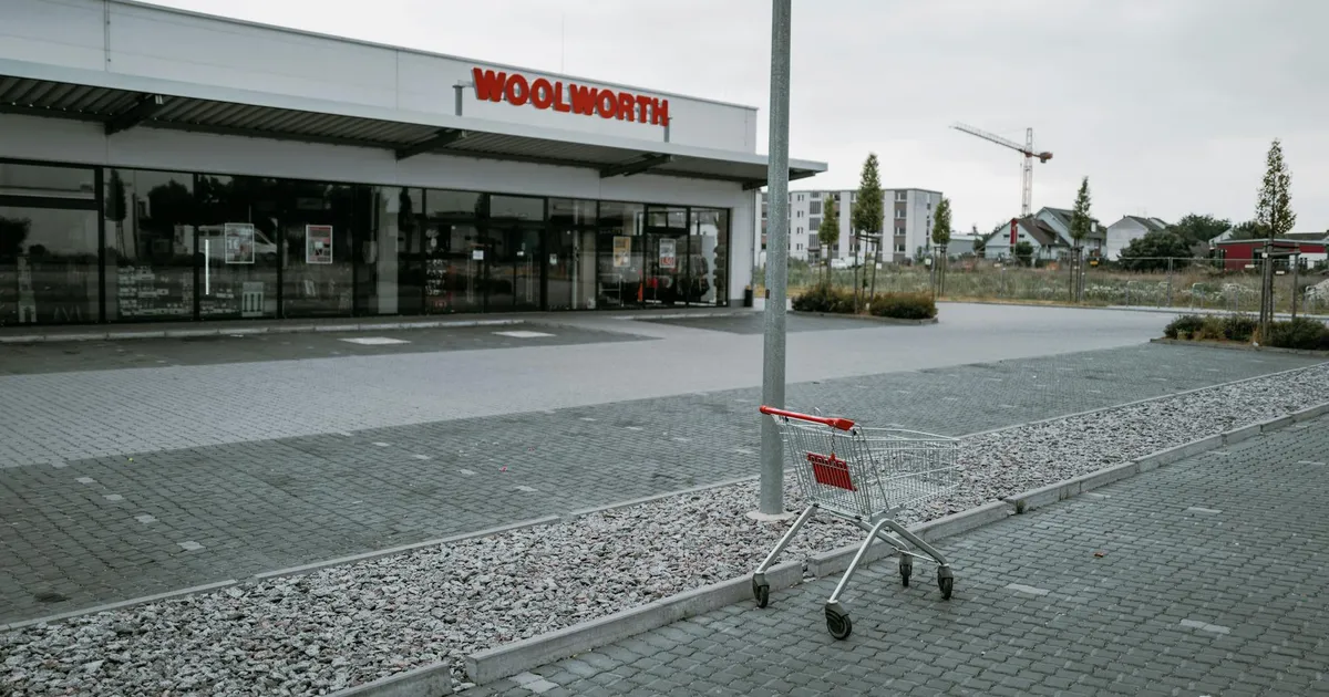 An empty parking lot with an abandoned shopping cart in front of a Woolworth store on a cloudy day.