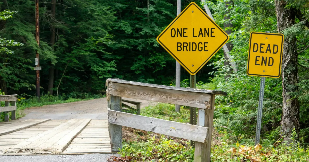 A rustic wooden one-lane bridge with 'Dead End' and 'One Lane Bridge' signs in a lush forest setting.