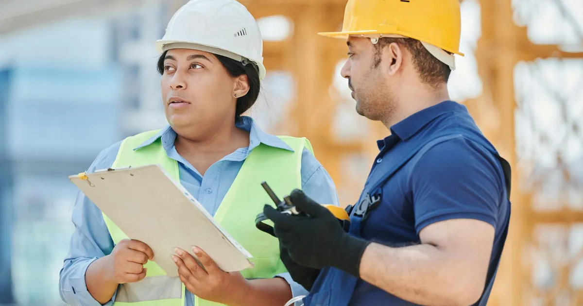 Male and female construction workers converse on a building site wearing hard hats.