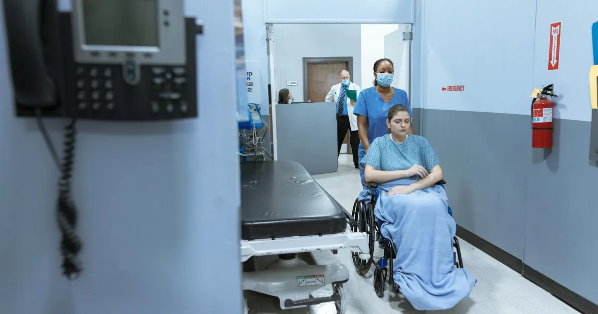 A nurse helps a patient in a wheelchair down a hospital corridor, reflecting care and medical professionalism.