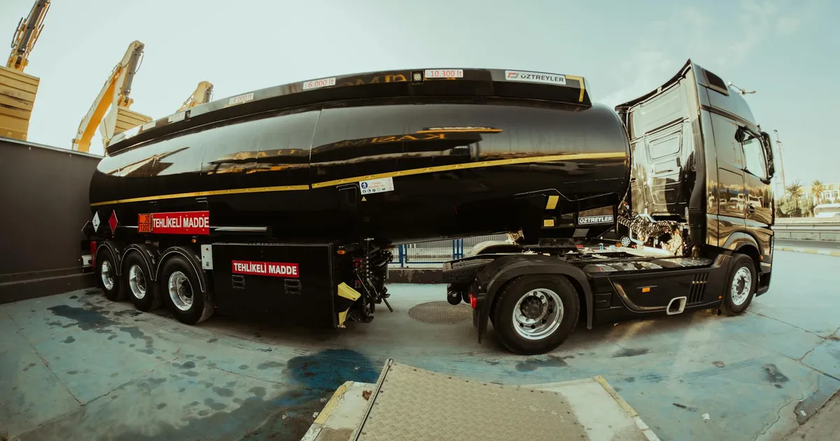 Side view of a black tanker truck parked at an industrial shipping facility under clear skies.