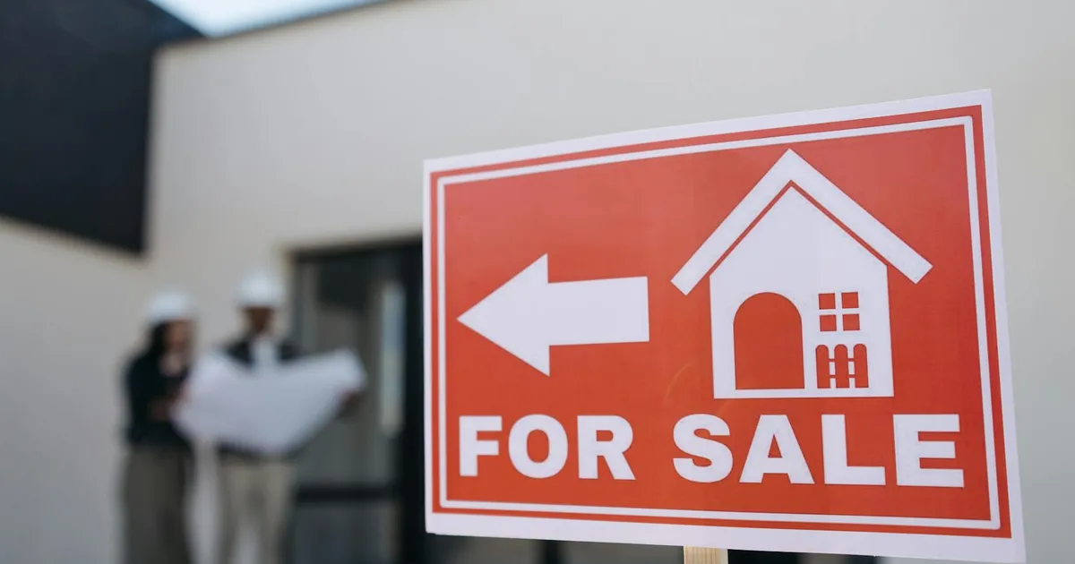 A real estate sign indicates a property for sale as two agents in hard hats discuss building plans outdoors.