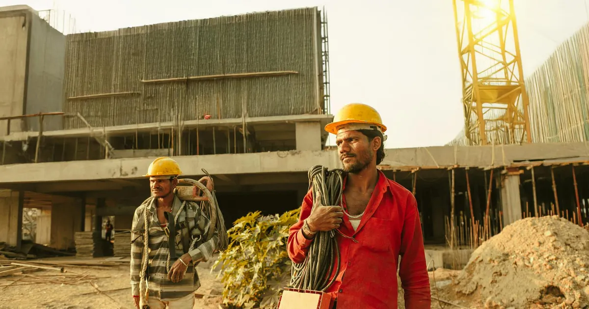 Two construction workers wearing helmets at a site in India with a crane and building.