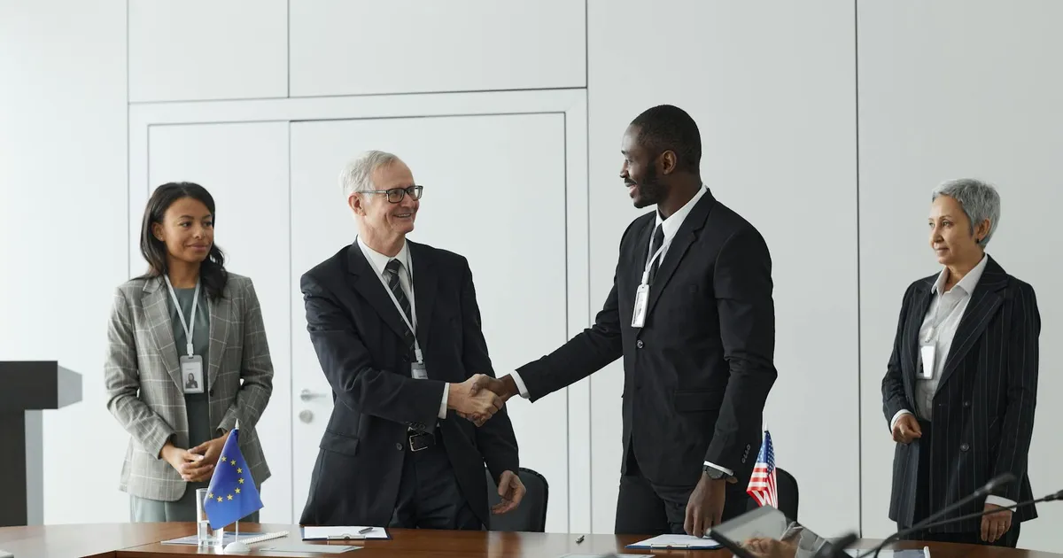 Business professionals shaking hands in conference room with flags signifying international agreement.
