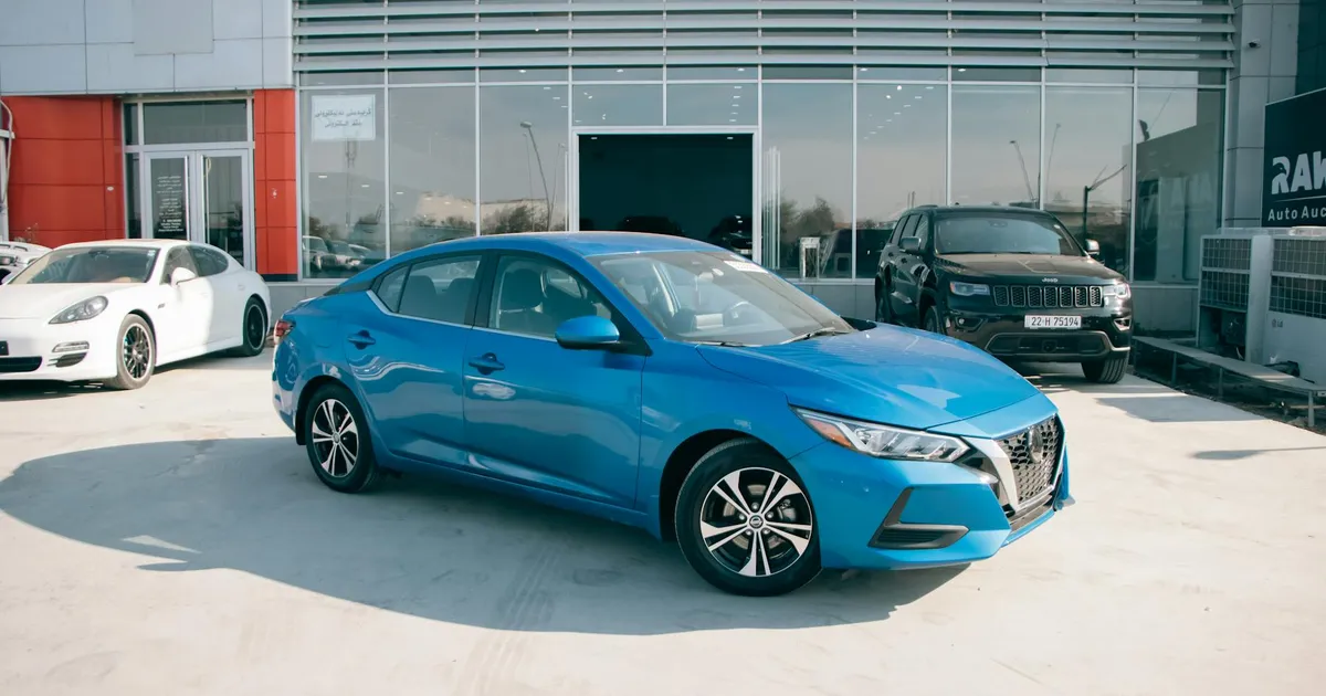 A bright blue Nissan Sentra parked in front of a car dealership on a sunny day.