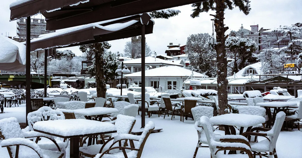 A winter scene of an outdoor restaurant covered in snow, with empty chairs and tables.