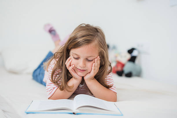 young girl lying on bed, resting on elbows and reading a book