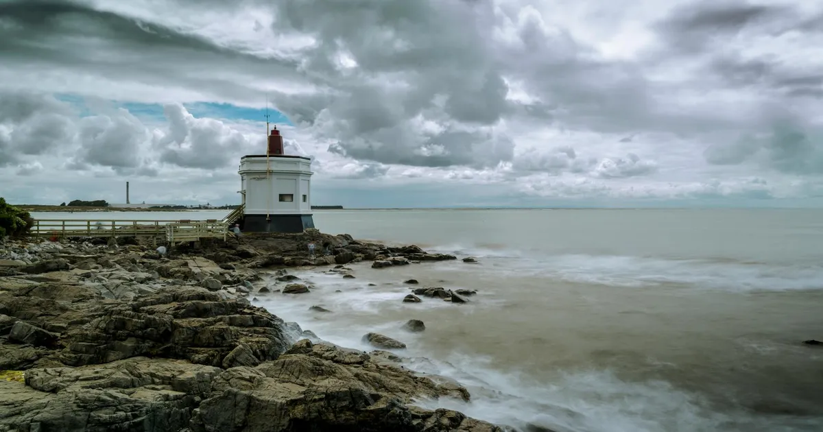 Lighthouse at Bluff, New Zealand with dramatic clouds over rocky shore and ocean.
