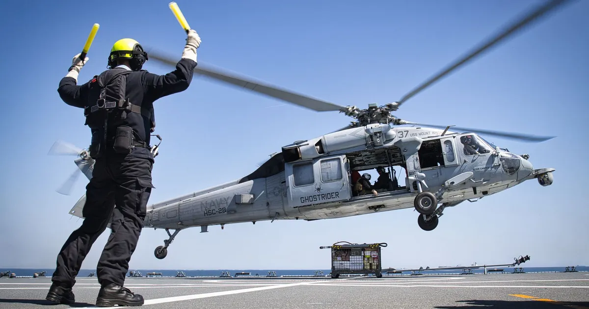 A military helicopter lands on an aircraft carrier guided by a deck crew member.