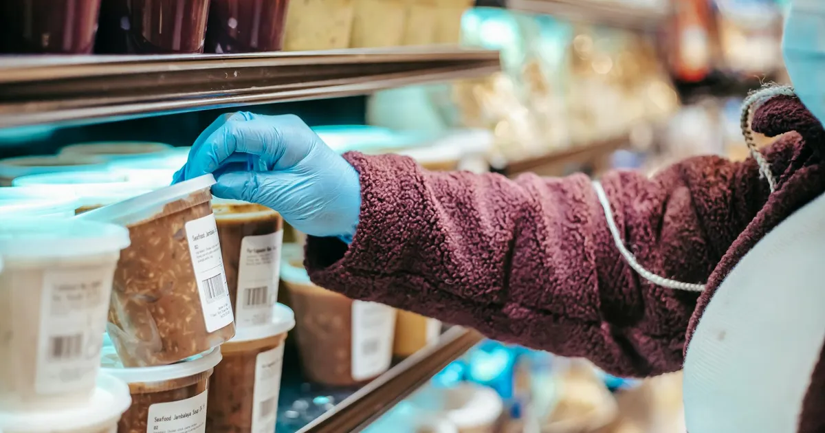Crop anonymous female customer in protective mask reading label on frozen food in plastic container in grocery store