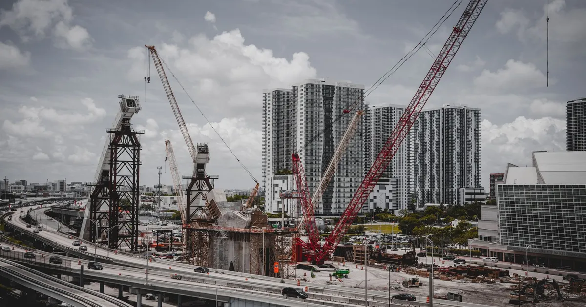 A cityscape featuring high-rise buildings and a construction site with cranes under a cloudy sky.