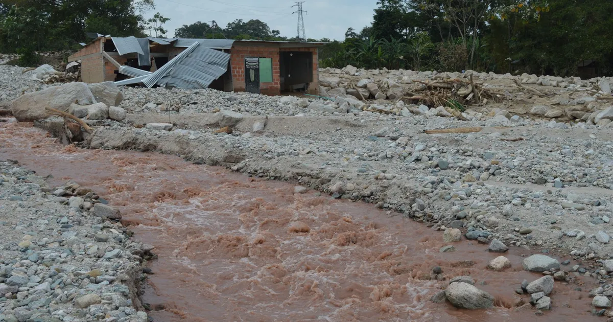A damaged house stands near a muddy river after a flood, illustrating the impact of natural disasters.
