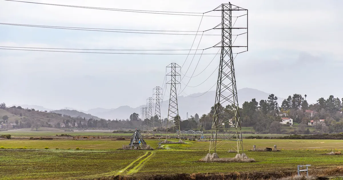 View of electric transmission towers stretching across a rural field with mountains in the background.