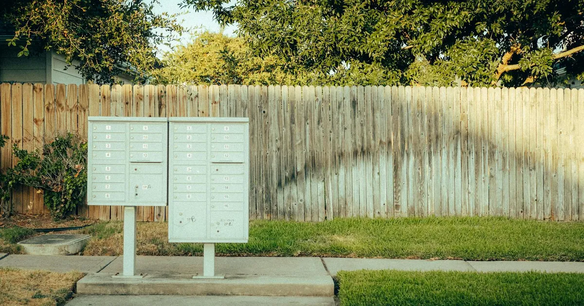 Mailboxes in a suburban area with a wooden fence and trees in the background.