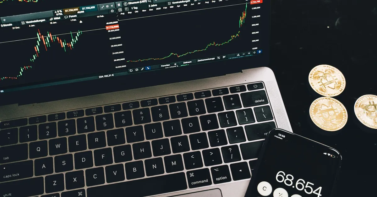 A laptop with trading charts, smartphone calculator, and bitcoin coins depicting cryptocurrency trading.