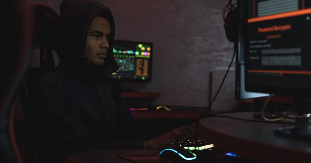 A hacker in a hoodie working in a dimly lit room, focusing on cyber security tasks on multiple monitors.