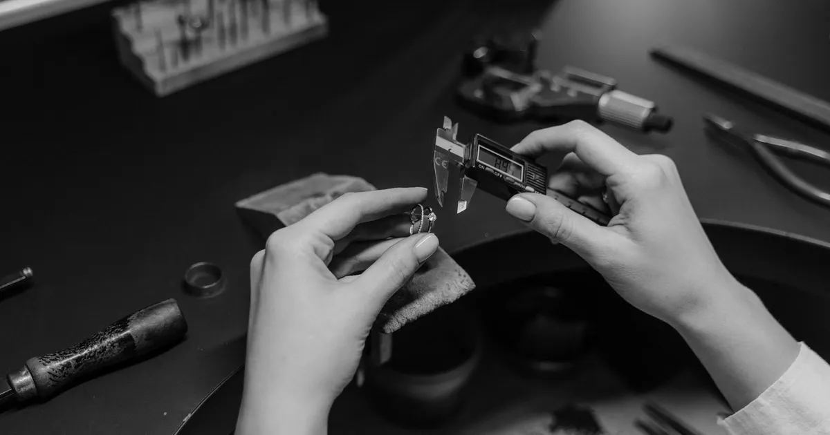 Black and white photo of a jeweler measuring a ring with a digital caliper in a workshop setting.