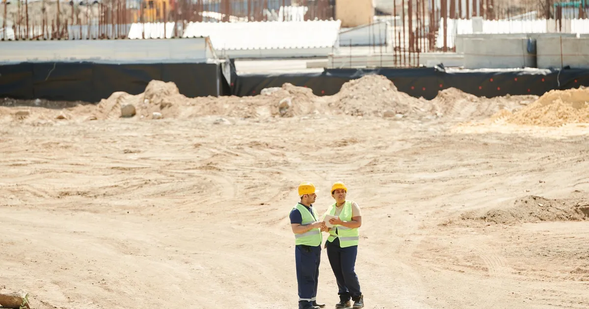 Two workers discussing plans on a sandy construction site, wearing safety gear.