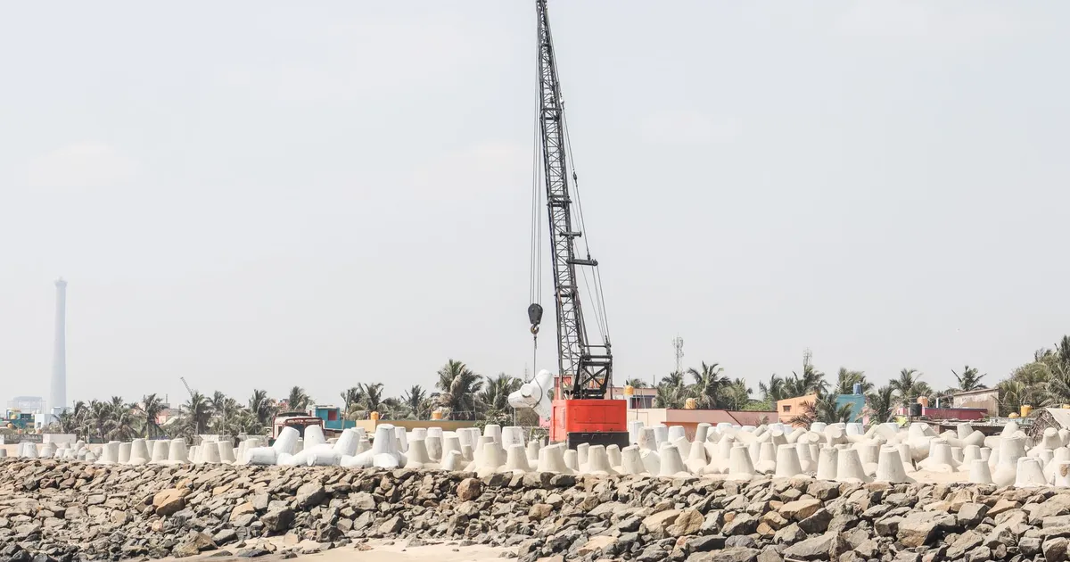 Crane constructing a seawall on a sandy beach with cement tetrapods and palm trees in the background.