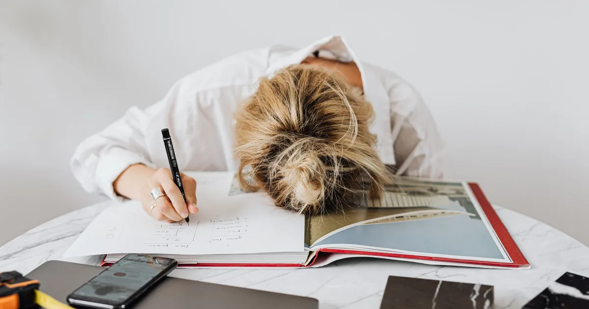 A tired architect rests head on open book at home office desk, indicating stress or fatigue.