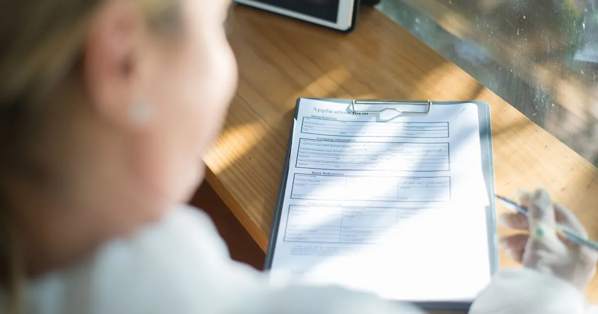 A woman fills out an application form with a pen at a sunlit wooden desk.