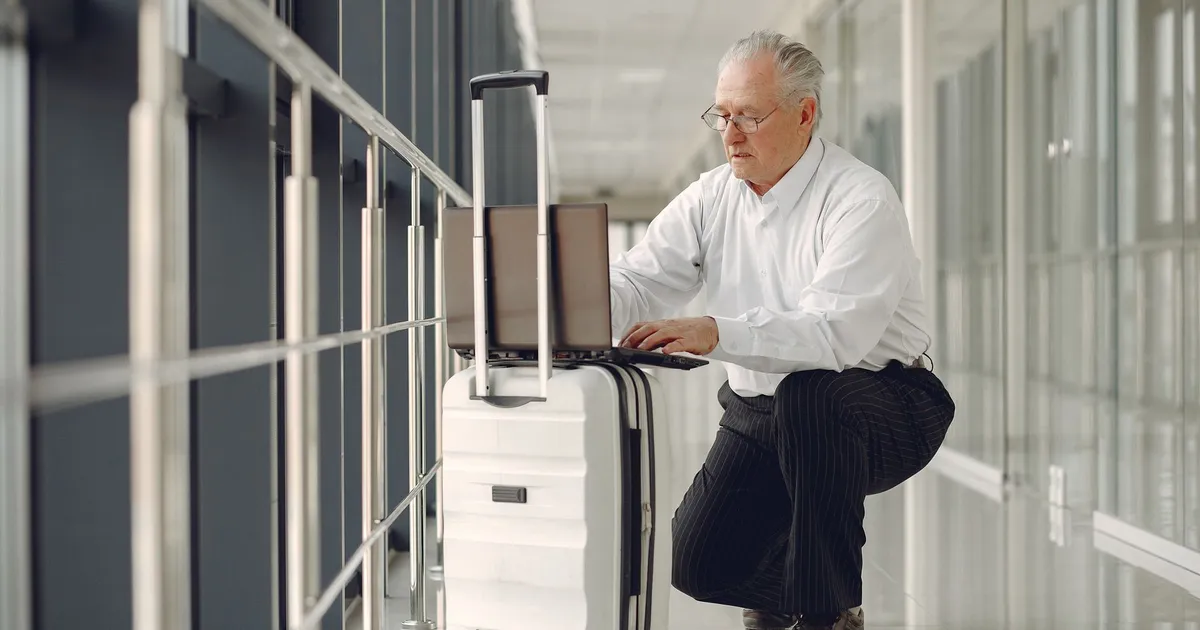 Full length of focused mature male in formal wear using laptop on baggage while having semisitting position in airport corridor