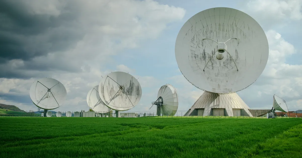 Array of large satellite dishes in a lush green field under a cloudy sky.