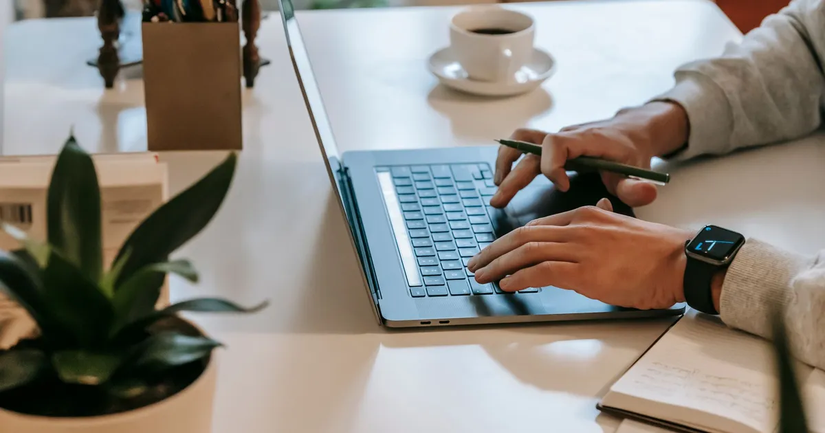 High angle side view of crop unrecognizable guy with smartwatch working remotely on laptop at table near cup of coffee on saucer near green potted plant and notebook in room