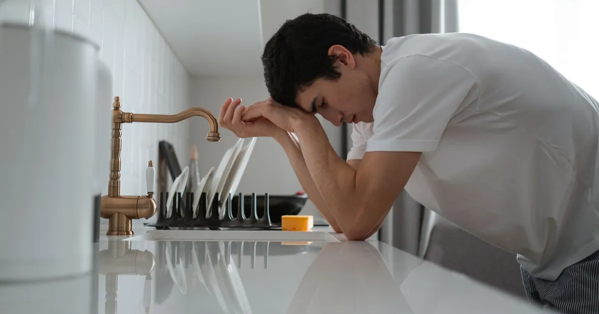 A young man appears tired and stressed, leaning on a kitchen counter in contemplation.