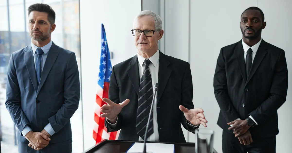 Three business professionals giving a speech with an American flag backdrop.