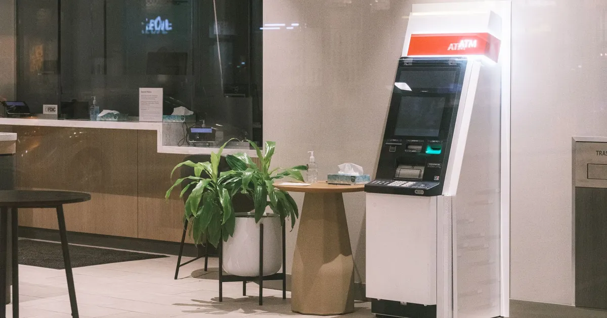 An ATM machine stands in a modern bank lobby, next to a plant and table.