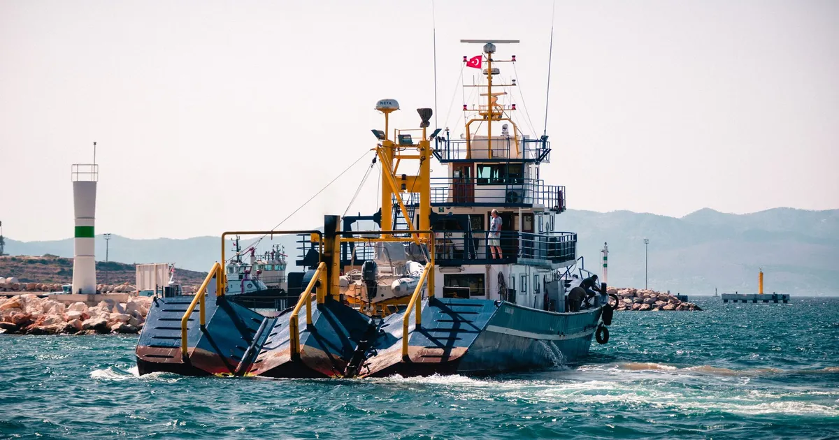 Large blue fishing boat with dredging equipment maneuvering in open waters near the coast.