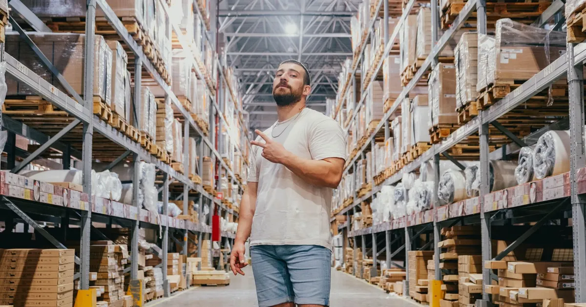 Young man posing in a warehouse aisle surrounded by racks and packages inside a spacious store.