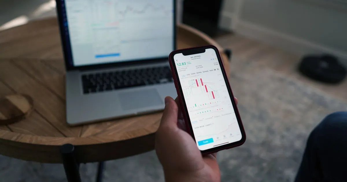 Person using smartphone with stock market charts while laptop displays data. Indoor setting.