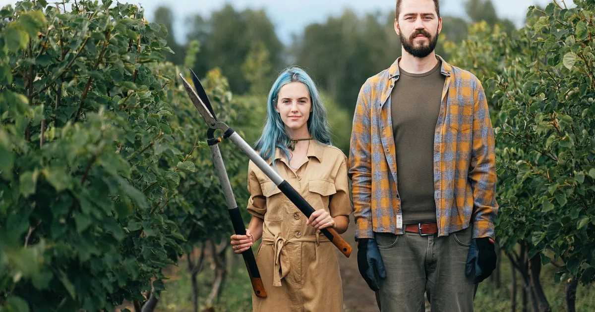 A young couple stands in a lush orchard, one holding gardening shears, symbolizing teamwork and organic farming.