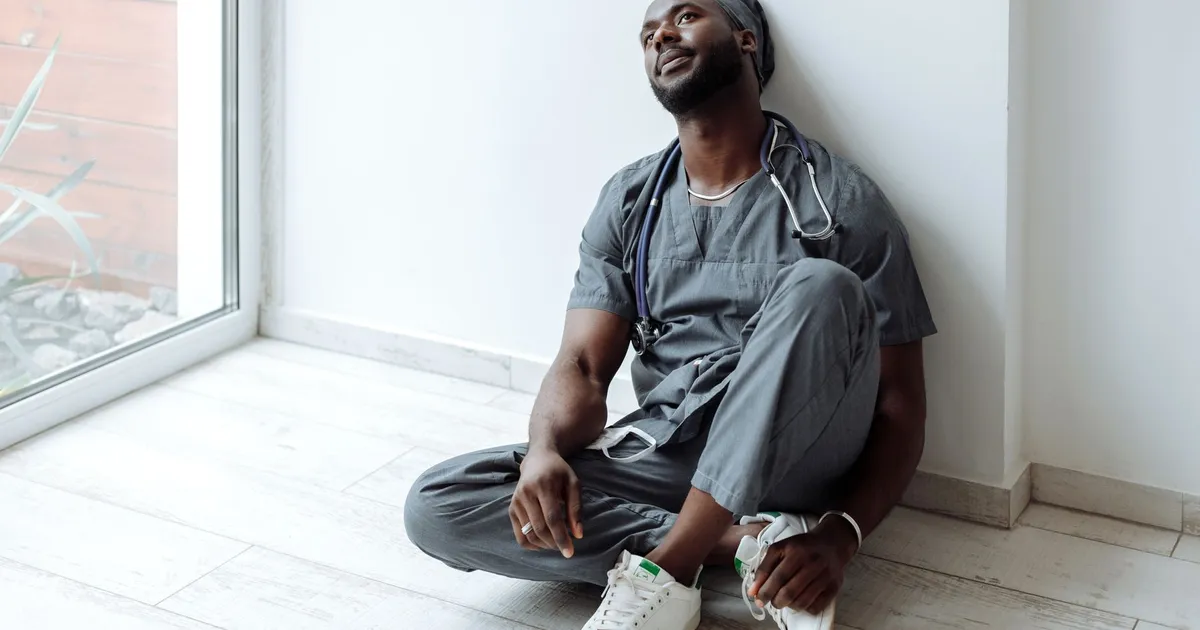 A tired healthcare professional in gray scrubs resting against a wall, reflecting exhaustion.