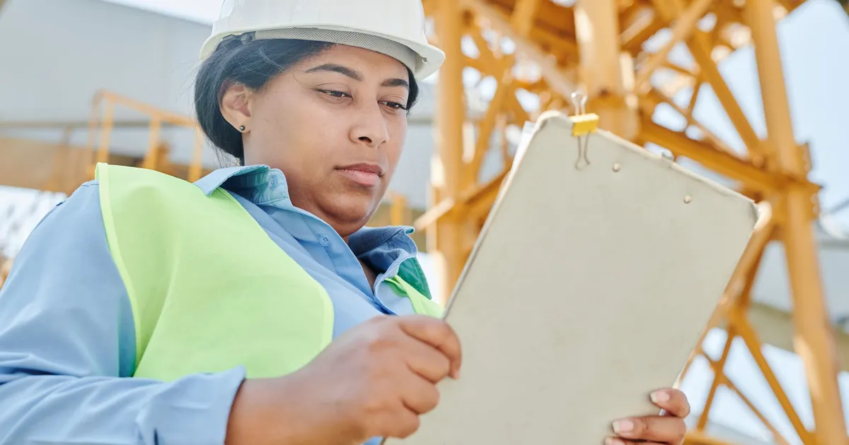 Confident female construction worker inspecting site with clipboard and helmet.